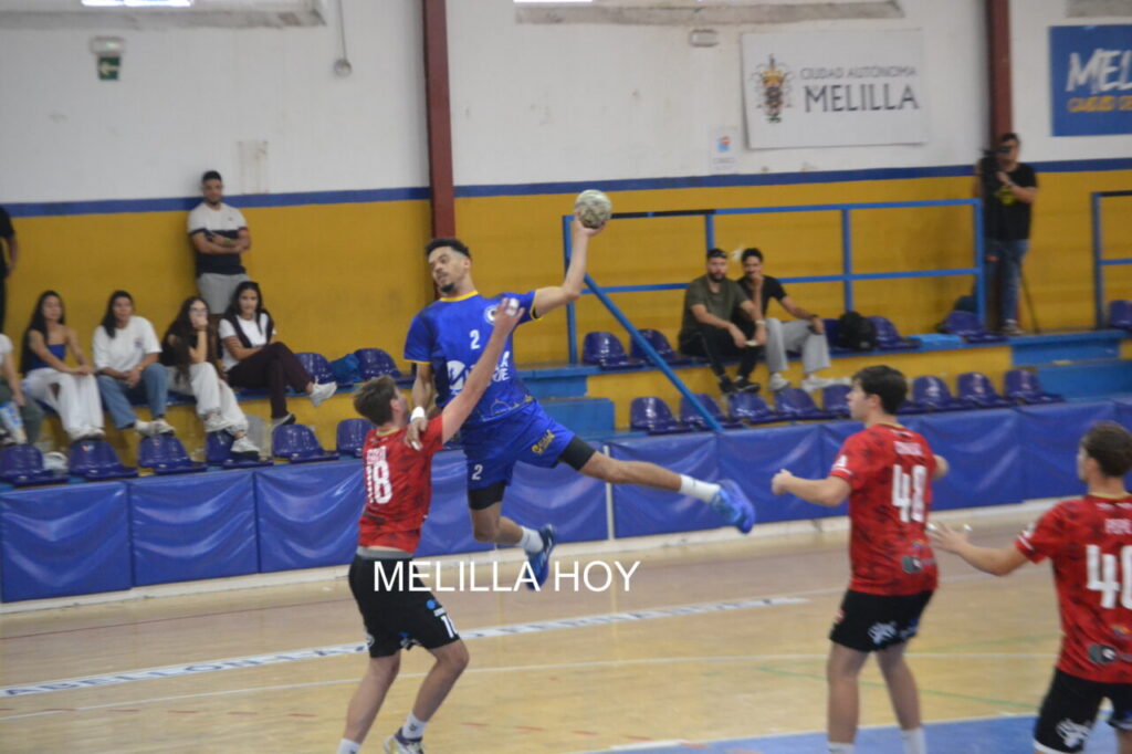 Jugador de azul salta y lanza el balón ante defensores de rojo en una cancha cubierta