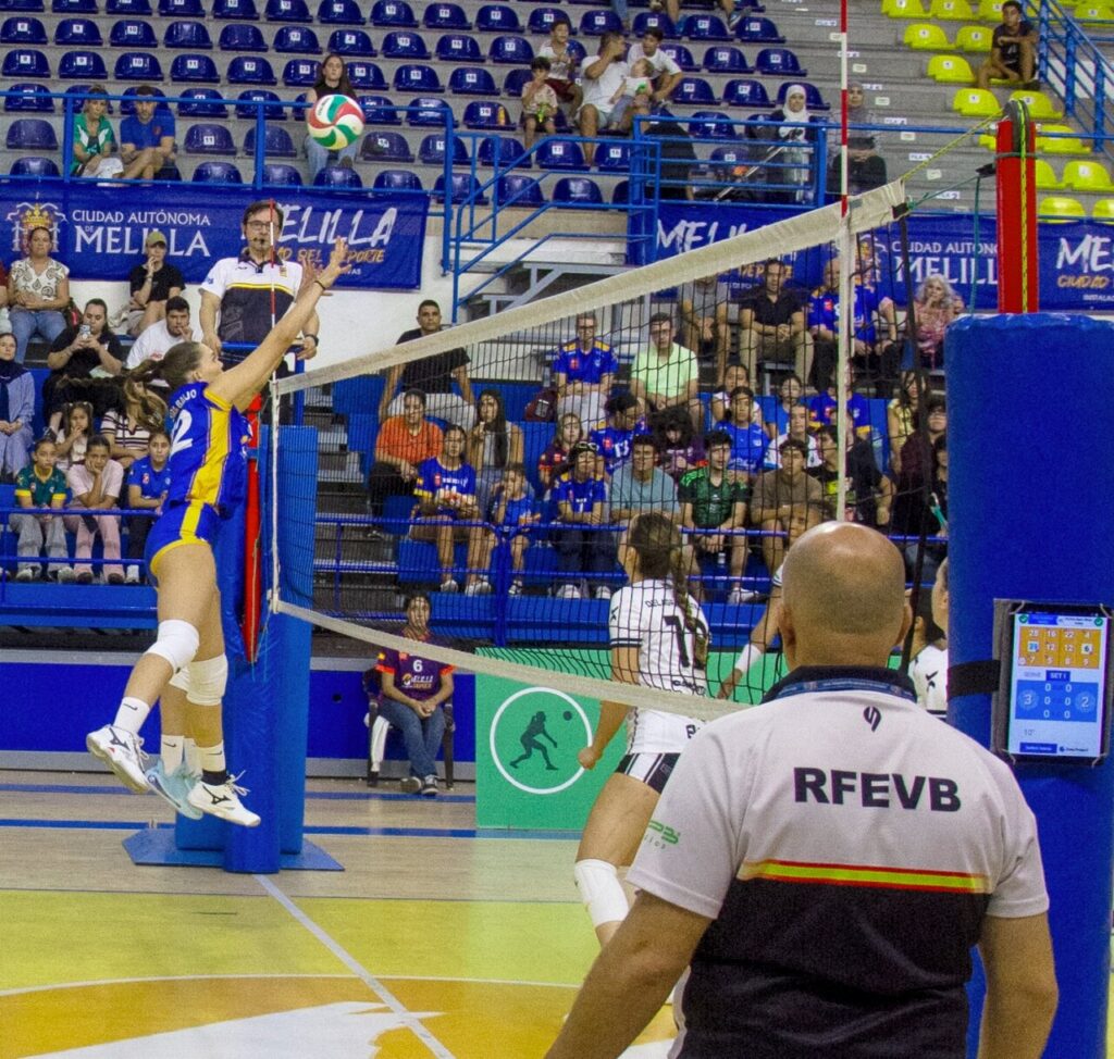Una jugadora salta junto a la red mientras el balón vuela sobre la pista.