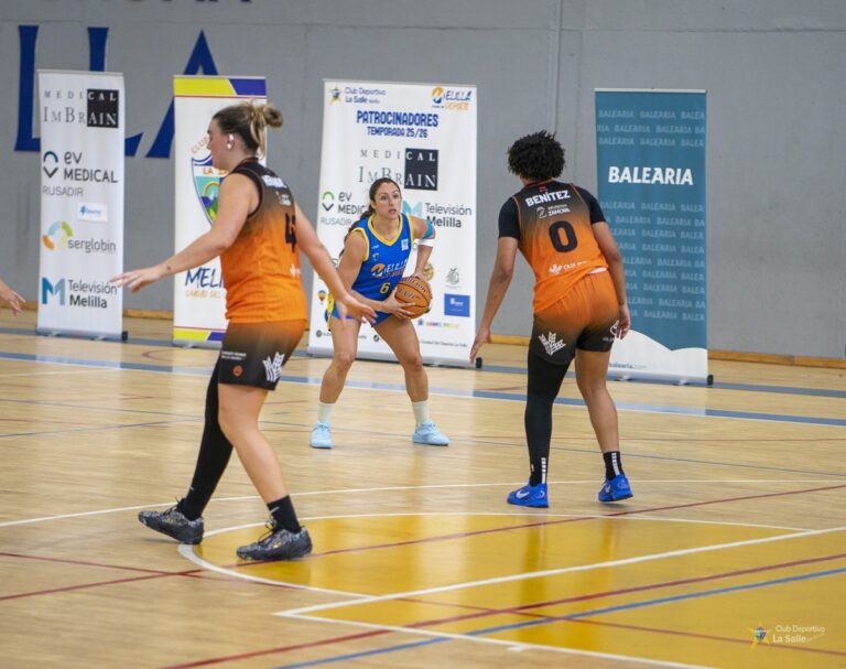 Una jugadora de azul sostiene el balón mientras dos rivales de naranja la defienden