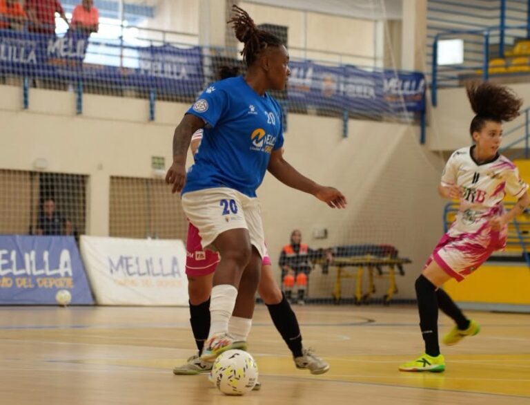 Una jugadora de azul controla el balón mientras otra se acerca en una pista cubierta.