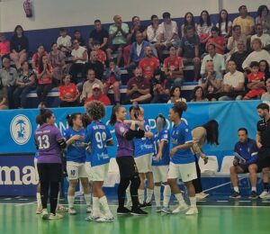 Female futsal players in blue uniforms exchange high-fives on the court as spectators watch from the stands behind them.