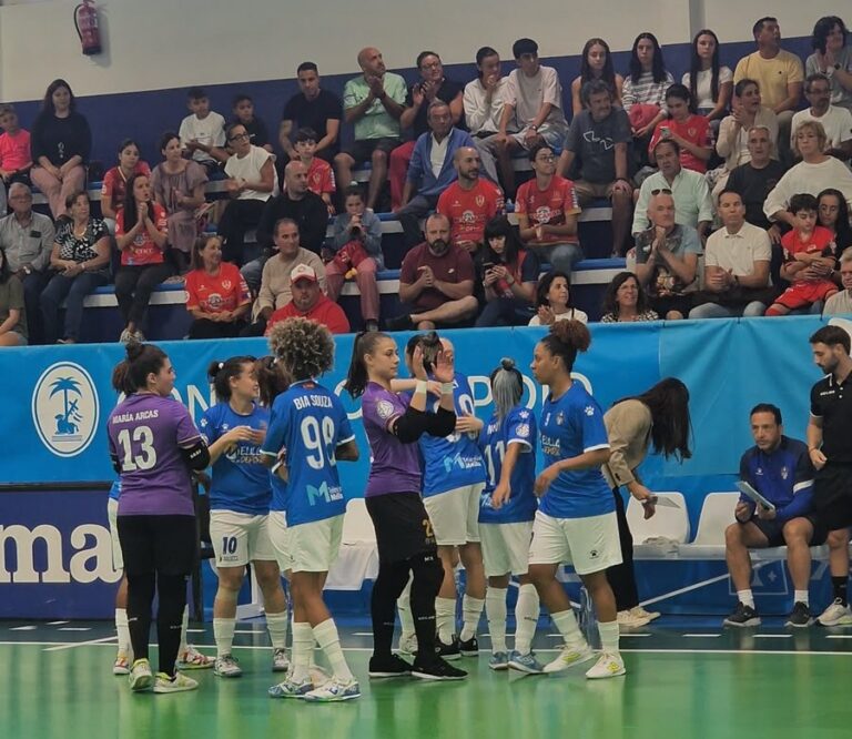 Female futsal players in blue uniforms exchange high-fives on the court as spectators watch from the stands behind them.