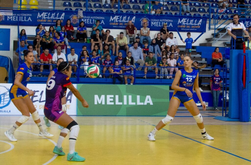 Action shot of a women's indoor volleyball match: players in blue and purple uniforms scramble for a ball as spectators watch from the stands with Melilla banners behind them.