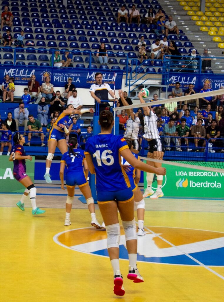 Volleyball match in a sports arena; players in blue and yellow uniforms jump near the net as one spikes the ball while defenders block; spectators sit in blue and yellow seats.