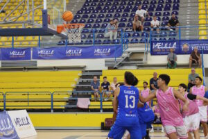 Jugadores de baloncesto en azul y rosa observan el balón junto a la canasta.