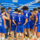 Jugadores del Enrique Soler, con uniformes azules y naranjas, se reúnen en círculo en la pista.