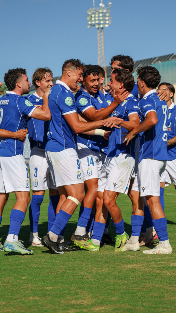Jugadores de la U.D. Melilla, con uniforme azul, se abrazan en el campo.