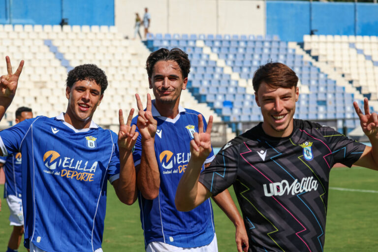Tres futbolistas con camisetas de la U.D. Melilla hacen gestos de victoria en el estadio