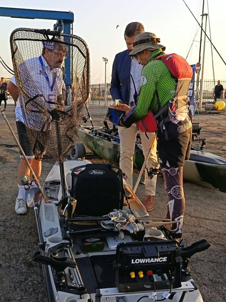 Three people on a dock with fishing kayaks and gear, one in a green suit showing a device to two others at sunset near a Lowrance unit on a kayak spurting fishing gear.