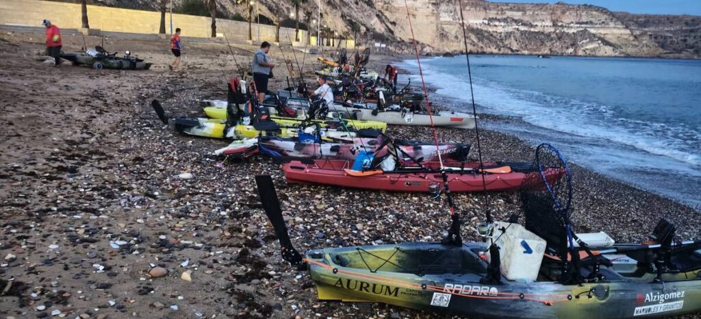 Colorful fishing kayaks and small boats lined up on a pebbly beach, with people preparing gear beside the shoreline at dusk.