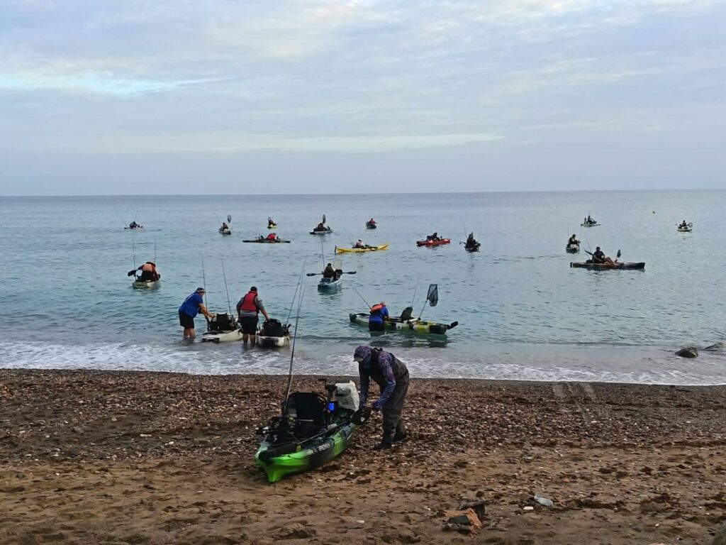 Personas en kayaks de pesca frente a la costa y una embarcación en la orilla