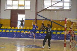 Jugador de balonmano lanza a portería mientras un portero intenta bloquear el tiro