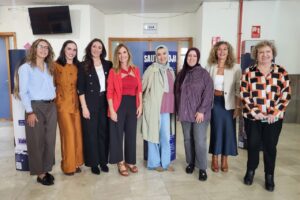 Eight women pose for a group photo indoors, dressed in business-casual outfits, smiling at the camera.