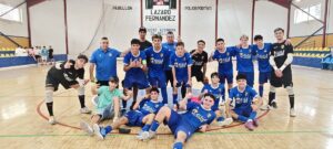 Group of young futsal players in blue uniforms pose for a team photo in an indoor gym, with two goalkeepers in black on either end and a coached standing behind.