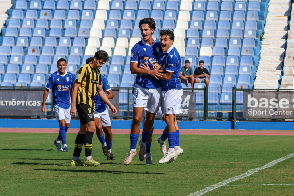 Soccer players in blue celebrate a goal, jumping and embracing on the field as an opposing player in black and yellow walks by with a disappointed expression.