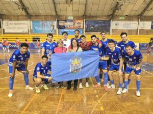 Grupo de jugadores con camisetas azules posa junto a una bandera en una pista cubierta.