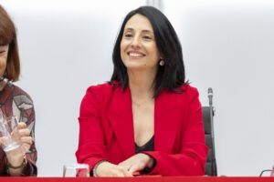 Mujer con blazer rojo sentada a una mesa junto a otra persona parcialmente visible