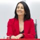 Mujer con blazer rojo sentada a una mesa junto a otra persona parcialmente visible