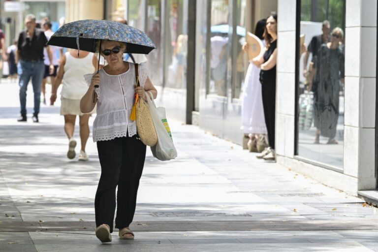 Mujer camina por una acera soleada mientras se protege con una sombrilla.