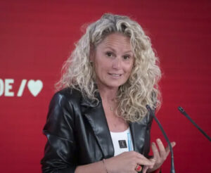 Mujer de cabello rizado habla ante dos micrófonos frente a un fondo rojo.