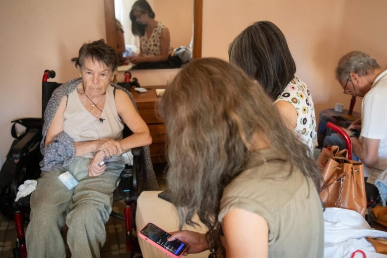 Mujer mayor sentada en una silla de ruedas, acompañada por varias personas en una habitación.