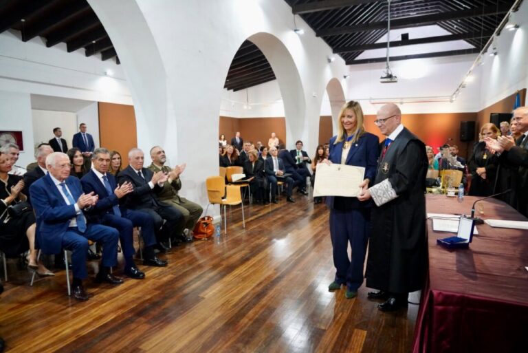 Una mujer sostiene un documento junto a un hombre con toga ante asistentes sentados.