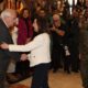 Woman in a white jacket shakes hands with an older man in a suit, as photographers and a soldier look on indoors at a ceremonial event with flags in the background.