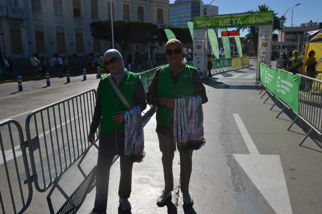 Dos mujeres con chalecos verdes sostienen medallas junto al arco de meta de una carrera.