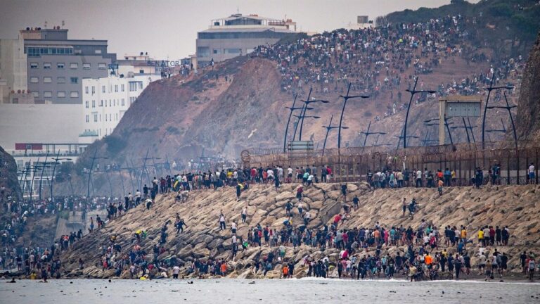 Multitud congregada en una costa rocosa junto a una valla y una ladera.