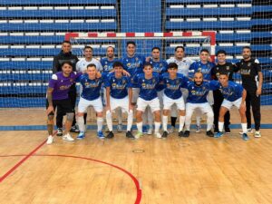 Group portrait of a futsal team in blue jerseys and white shorts posing with coaches in black, inside a gym with a goal behind.