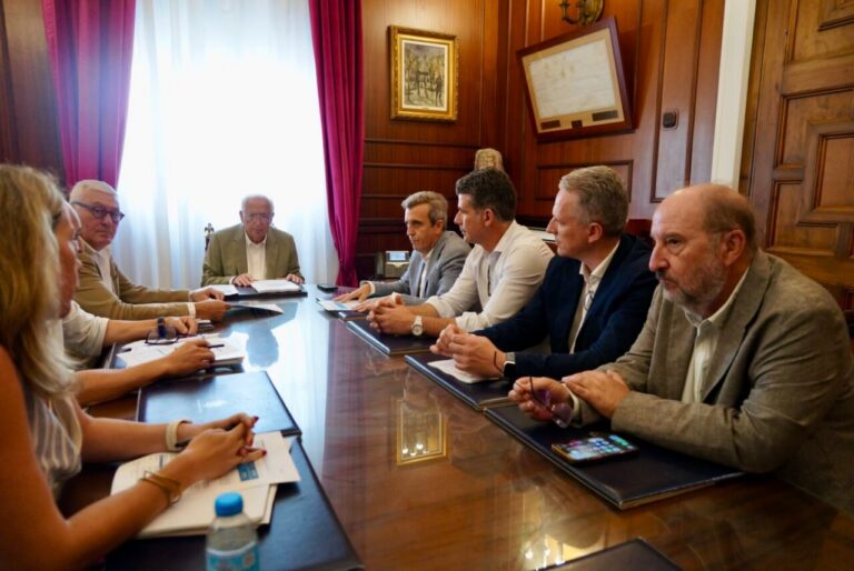 A group of professionals sit around a long wooden conference table in a formal meeting room, papers and notebooks in front of them as they discuss.