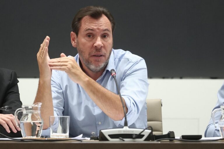 Man in a light blue shirt speaks into a microphone at a panel, using a hand gesture at the table.