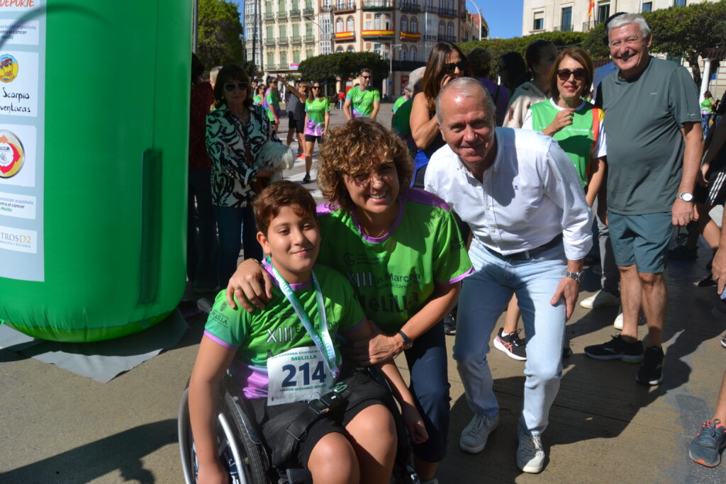 Participante en silla de ruedas posa junto a dos adultos durante una carrera popular en Melilla.