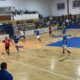 Soccer/futsal match indoors with players in red and blue; red player dribbles ball toward goal as blue players defend.