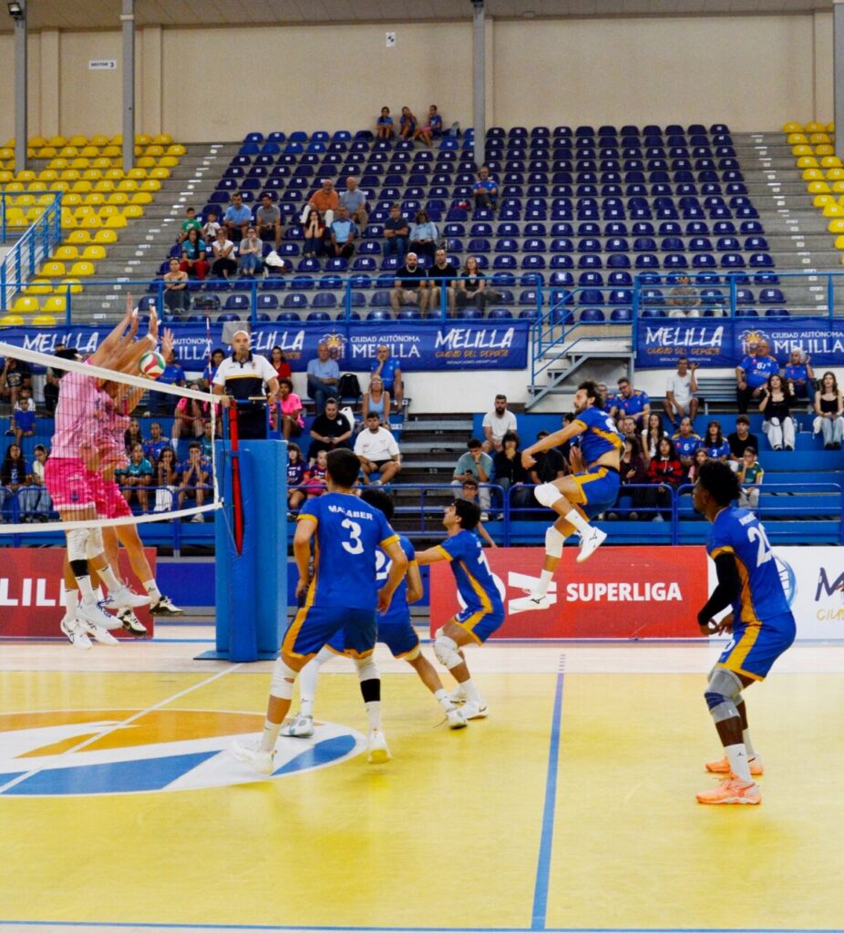 Jugadores disputan un punto de voleibol junto a la red mientras el público observa.