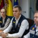 Pedro Sánchez sentado en una mesa junto a otras personas, con banderas al fondo.