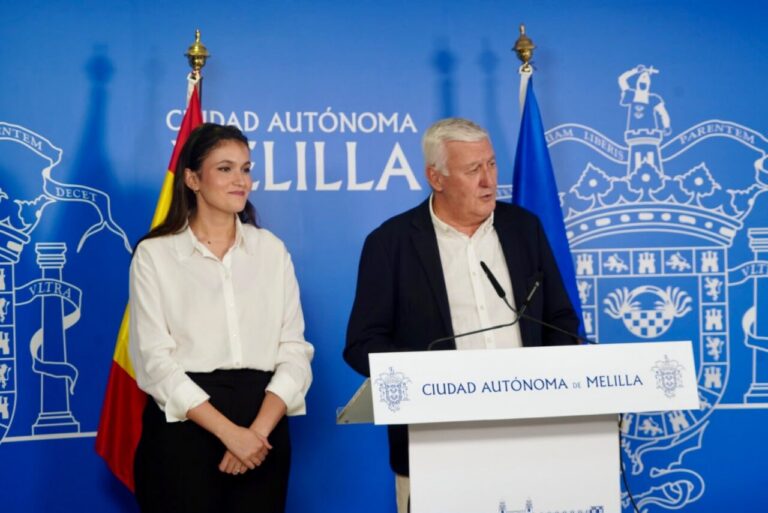 Two presenters stand at a white podium with microphones, a woman in a white blouse and black skirt beside a man in a white shirt and dark blazer, against a blue Melilla government backdrop and flags.