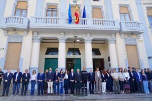 Personas alineadas frente a un edificio con columnas y varias banderas en la fachada