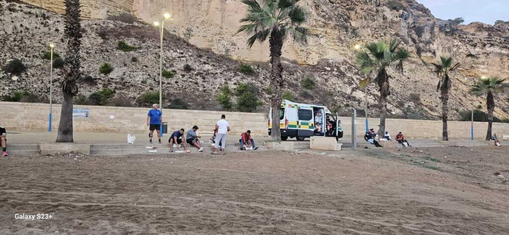 Personas reunidas en una playa bajo palmeras, junto a una furgoneta y acantilados.