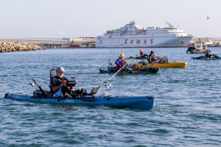 Varias personas pescan desde kayaks en el puerto, con un ferry al fondo.