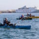 Varias personas pescan desde kayaks en el puerto, con un ferry al fondo.