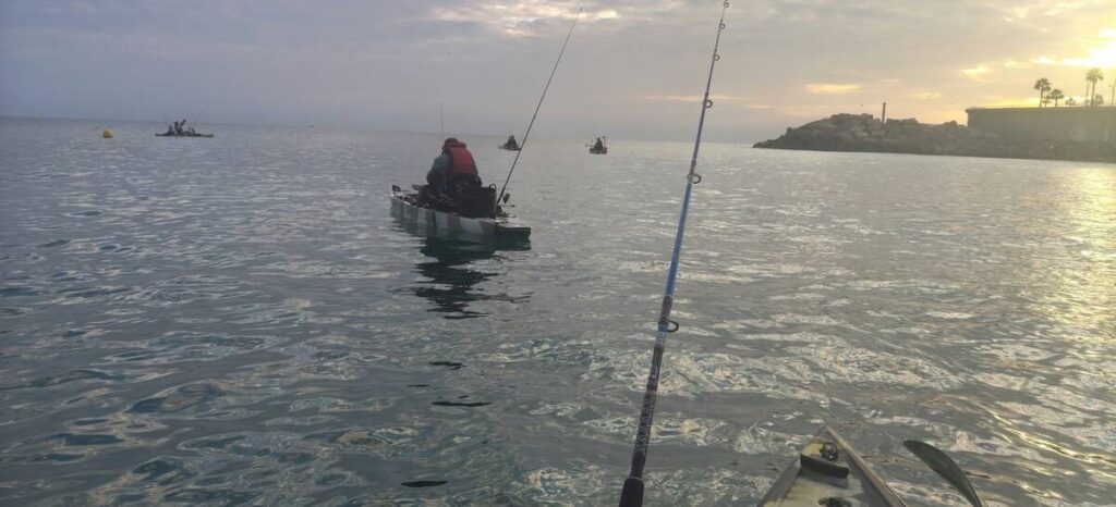 Varias personas pescan desde kayaks en el mar al atardecer, con una caña en primer plano.