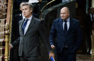 Two men in dark suits exit a grand stairwell, one with gray hair and blue tie, the other bald in a navy suit.