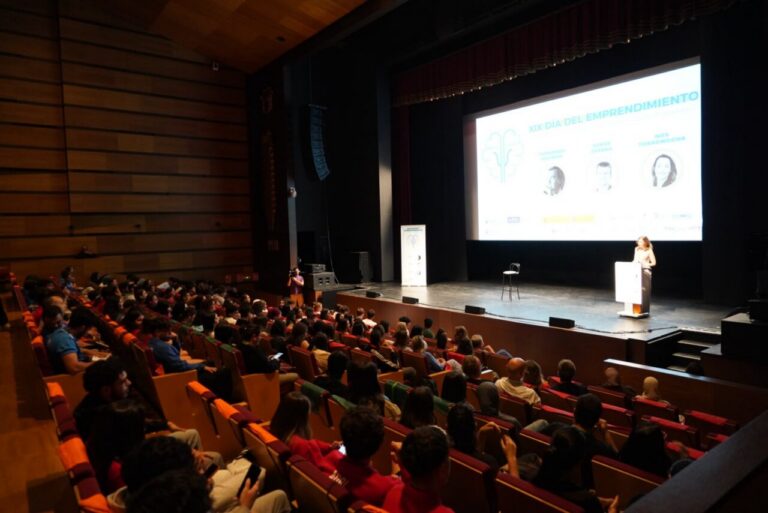 Audience in an auditorium watching a presenter on stage with a large screen showing 'XIX Día del Emprendimiento'.