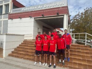 Group of young male athletes in red jerseys posing on brown tiled steps in front of a sports pavilion labeled Pabellón de Deportes.