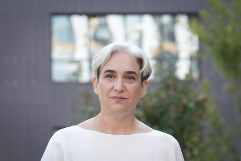 Retrato de Ada Colau con blusa blanca ante un fondo urbano desenfocado