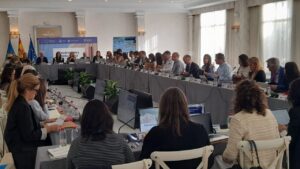 Large conference room with a long U‑shaped table of attendees, laptops, and microphones during a formal economic forum.