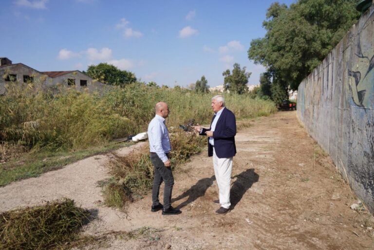 Dos hombres conversan en un sendero junto a vegetación alta y una pared pintada.