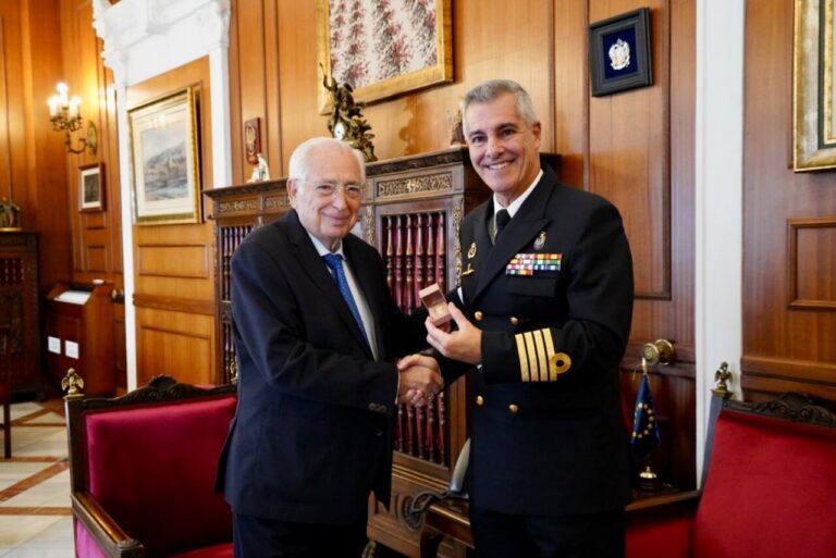 Dos hombres se estrechan la mano; uno viste uniforme naval y sostiene una pequeña caja.