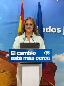 Woman at a podium delivering a speech, with a blue banner reading 'El cambio está más cerca' and flags in the background.